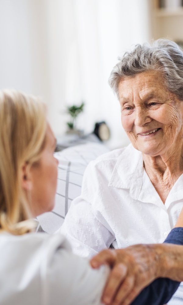 A health visitor talking to a sick senior woman sitting on bed at home.